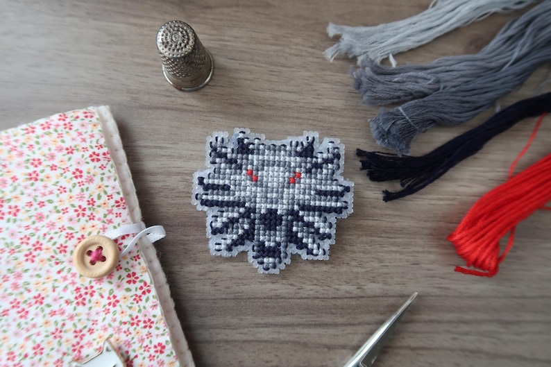 May include: A cross-stitch patch featuring a wolf's head design with red eyes, in shades of gray and black. The patch is surrounded by embroidery floss in gray, black, and red, a thimble, a needle, and a floral fabric-covered notebook.
