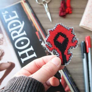 May include: A red and black cross-stitch emblem, held in a hand, featuring a stylized design. The emblem is in the shape of a stylized symbol. The word "HORDE" is visible in the background.