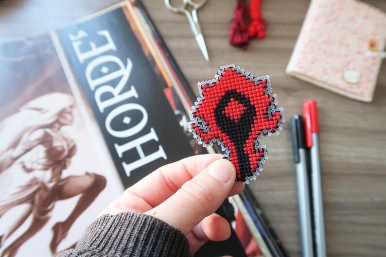 May include: A red and black cross-stitch emblem, held in a hand, featuring a stylized design. The emblem is in the shape of a stylized symbol. The word "HORDE" is visible in the background.