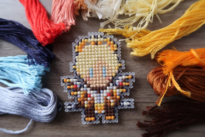 May include: A cross-stitch design of a character with blonde hair, blue eyes, and a brown and orange outfit. The design is surrounded by colorful embroidery floss in shades of red, pink, blue, yellow, and brown. The craft supplies are arranged on a wooden surface.