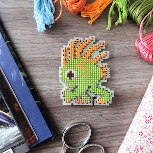 May include: A pixelated green creature cross-stitch design with orange accents. The image includes colorful embroidery floss, fabric, and scissors, suggesting a crafting theme. The design is on a wooden surface.