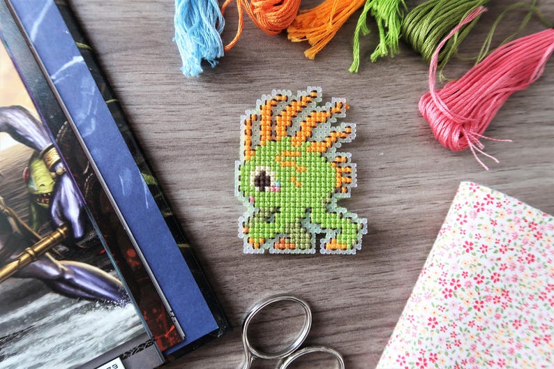 May include: A pixelated green creature cross-stitch design with orange accents. The image includes colorful embroidery floss, fabric, and scissors, suggesting a crafting theme. The design is on a wooden surface.
