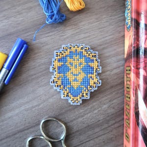 May include: A cross-stitched emblem in blue and gold, featuring a detailed design. The emblem is surrounded by craft supplies, including blue and yellow markers, scissors, and thread. A book with a red spine is also visible.