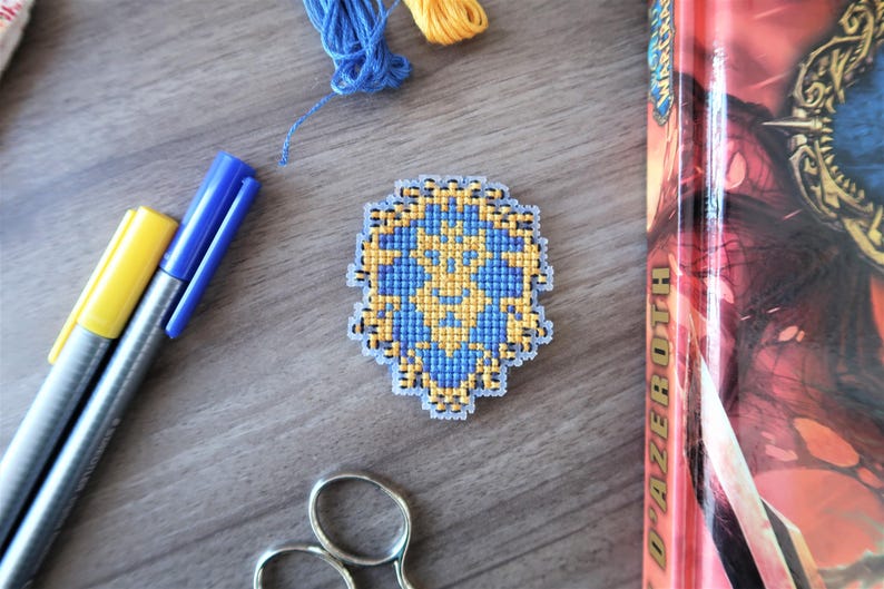 May include: A cross-stitched emblem in blue and gold, featuring a detailed design. The emblem is surrounded by craft supplies, including blue and yellow markers, scissors, and thread. A book with a red spine is also visible.