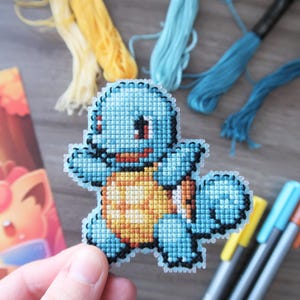 May include: A cross-stitch of a blue Squirtle character with a yellow and orange shell. The image includes spools of yellow and blue thread, colored markers, and a postcard with a cartoon character.
