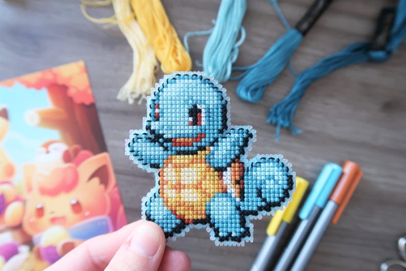 May include: A cross-stitch of a blue Squirtle character with a yellow and orange shell. The image includes spools of yellow and blue thread, colored markers, and a postcard with a cartoon character.