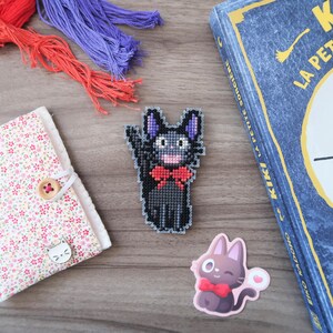 May include: A collection of craft supplies and Kiki's Delivery Service themed items. Includes a floral fabric-covered notebook, a cross-stitched black cat with a red bow tie, a sticker of a cat with a bow tie, embroidery floss, and a book.