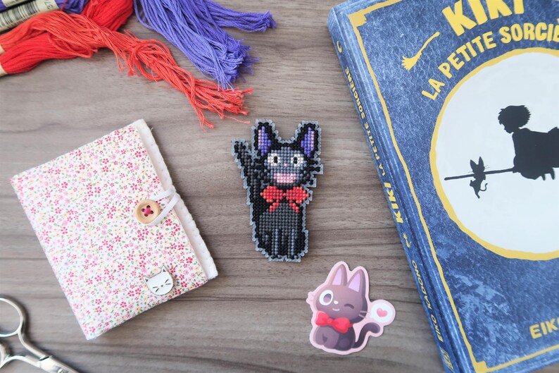 May include: A collection of craft supplies and Kiki's Delivery Service themed items. Includes a floral fabric-covered notebook, a cross-stitched black cat with a red bow tie, a sticker of a cat with a bow tie, embroidery floss, and a book.
