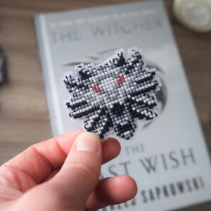 May include: A handmade, cross-stitched Witcher medallion in black, white, and gray, with red eyes. The medallion is held in front of a book titled "The Witcher: The Last Wish" by Andrzej Sapkowski.