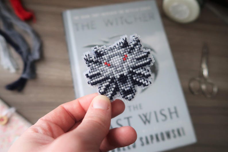 May include: A handmade, cross-stitched Witcher medallion in black, white, and gray, with red eyes. The medallion is held in front of a book titled "The Witcher: The Last Wish" by Andrzej Sapkowski.