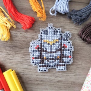 May include: A cross-stitch project featuring a pixelated robot design in shades of gray, brown, and yellow. Colorful embroidery floss in yellow, red, orange, gray, and brown surrounds the project. Markers and a floral fabric are also present.