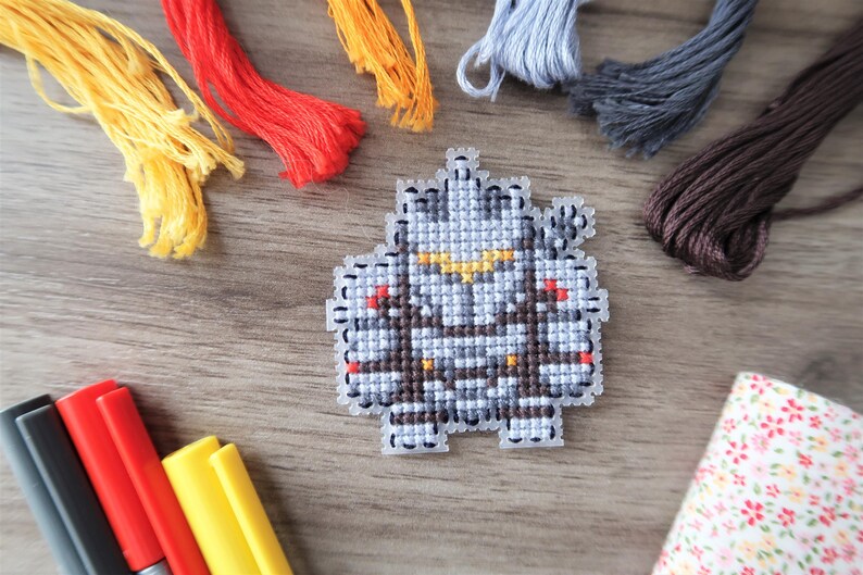May include: A cross-stitch project featuring a pixelated robot design in shades of gray, brown, and yellow. Colorful embroidery floss in yellow, red, orange, gray, and brown surrounds the project. Markers and a floral fabric are also present.