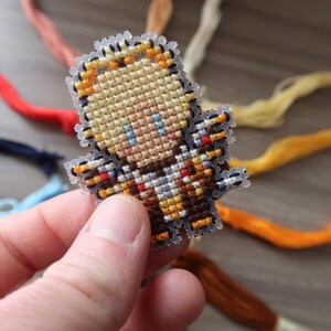 May include: A small, pixelated character made with cross-stitch embroidery. The character has blonde hair, blue eyes, and is wearing a brown and tan outfit. The edges are clear plastic. Colorful embroidery floss is in the background.