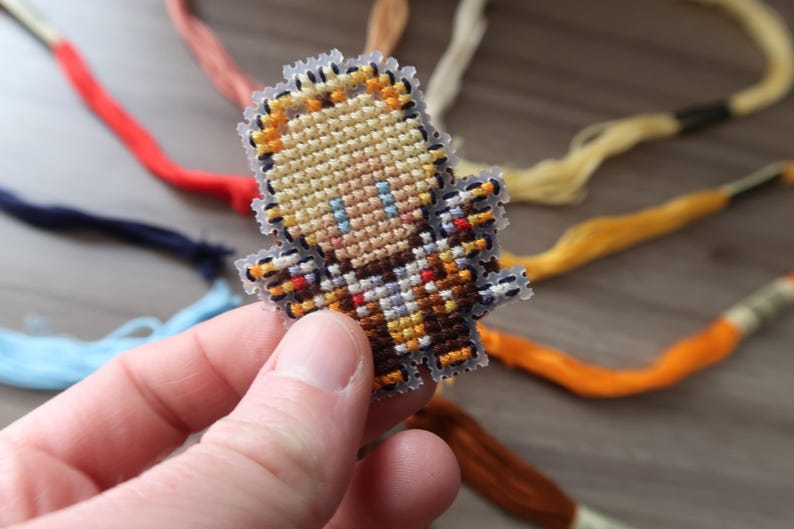 May include: A small, pixelated character made with cross-stitch embroidery. The character has blonde hair, blue eyes, and is wearing a brown and tan outfit. The edges are clear plastic. Colorful embroidery floss is in the background.