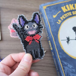 May include: A cross-stitch cat with a red bow tie, black body, and purple ears. The cat is waving. A book with the title "KIKI LA PETITE SORCIERE" is in the background. The book has a blue cover with yellow text.