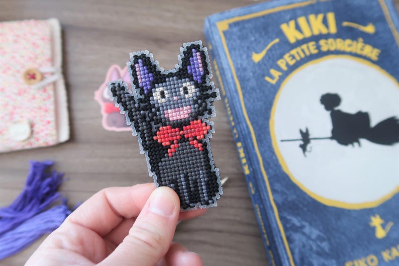 May include: A cross-stitch cat with a red bow tie, black body, and purple ears. The cat is waving. A book with the title "KIKI LA PETITE SORCIERE" is in the background. The book has a blue cover with yellow text.