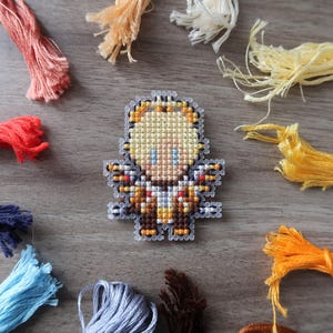 May include: A cross-stitch angel pin with yellow, brown, and blue details. The pin is surrounded by colorful embroidery floss in shades of red, orange, yellow, blue, and brown. The craft supplies are arranged on a wooden surface.