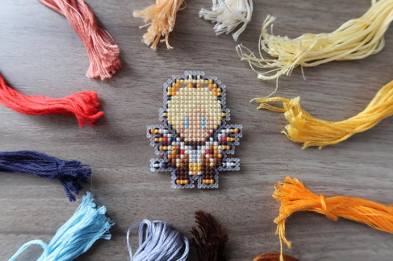 May include: A cross-stitch angel pin with yellow, brown, and blue details. The pin is surrounded by colorful embroidery floss in shades of red, orange, yellow, blue, and brown. The craft supplies are arranged on a wooden surface.