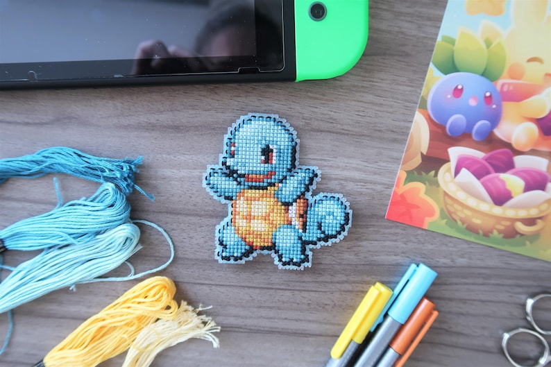 May include: A cross-stitched Squirtle patch in shades of blue and orange, with a detailed pixelated design. The patch is placed on a wooden surface, alongside embroidery floss, markers, and a Nintendo Switch.