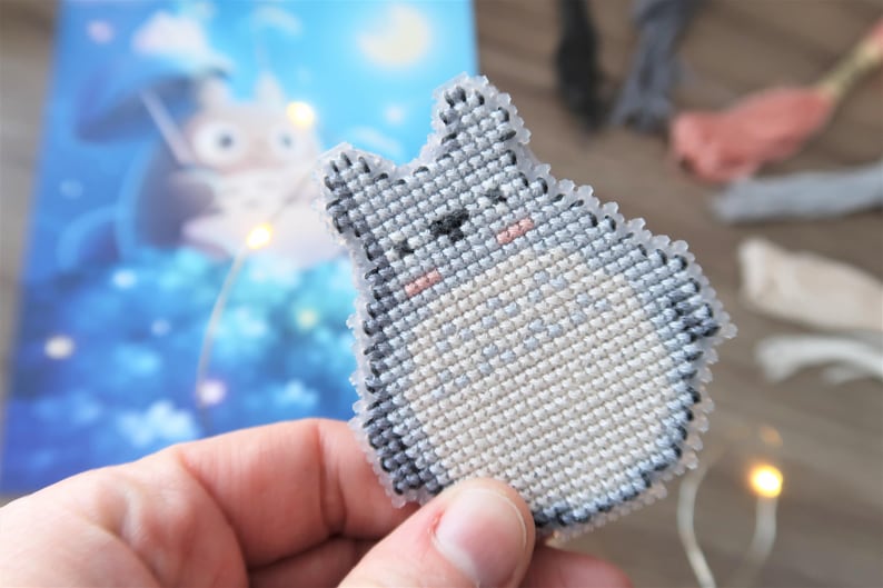 May include: A handmade cross-stitch of a Totoro character from the animated film, My Neighbor Totoro. The gray and white figure has a black nose and eyes, and pink cheeks. The cross-stitch is held in front of a blue and white background with a Totoro illustration.