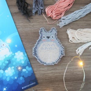 May include: A cross-stitch of a gray Totoro character with a white belly, surrounded by embroidery floss in various colors. A blue illustration with a moon and stars is on the left. A string of fairy lights is also present.