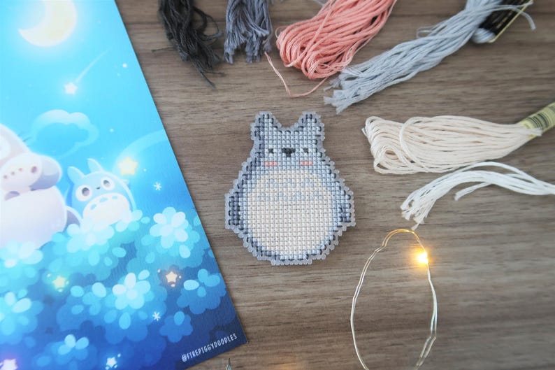 May include: A cross-stitch of a gray Totoro character with a white belly, surrounded by embroidery floss in various colors. A blue illustration with a moon and stars is on the left. A string of fairy lights is also present.