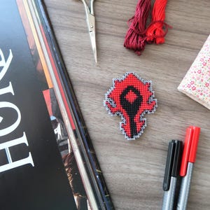 May include: A cross-stitched red and black emblem, possibly a game logo, sits on a wooden surface. Other craft supplies include red embroidery floss, a floral-patterned notebook, scissors, and markers. A magazine with the word "HORDE" is also present.