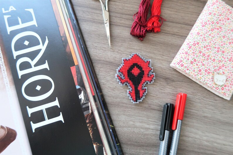 May include: A cross-stitched red and black emblem, possibly a game logo, sits on a wooden surface. Other craft supplies include red embroidery floss, a floral-patterned notebook, scissors, and markers. A magazine with the word "HORDE" is also present.