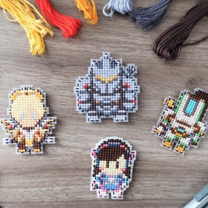 May include: A collection of pixel art character designs, created with cross-stitch techniques. The characters are in various colors and designs, with embroidery floss and craft supplies in the background. Scissors and markers are also visible.