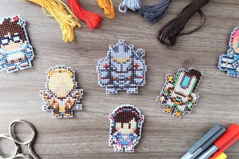 May include: A collection of pixel art character designs, created with cross-stitch techniques. The characters are in various colors and designs, with embroidery floss and craft supplies in the background. Scissors and markers are also visible.