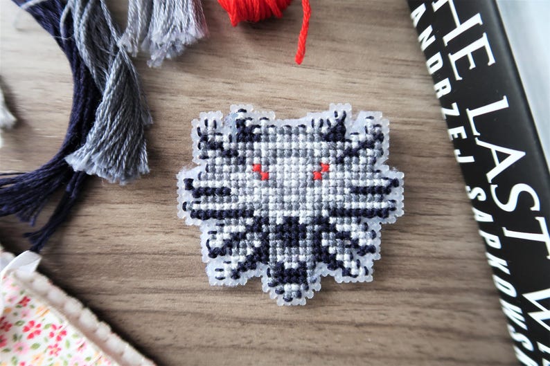 May include: A cross-stitch design of a wolf's head with red eyes, in shades of gray and black. The design is on a light-colored plastic canvas. Yarn tassels and a book are in the background.