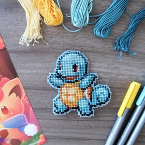 May include: A handmade cross-stitch of a blue cartoon turtle with a yellow belly, surrounded by embroidery floss and colorful markers. A colorful illustration of a fox is also present.