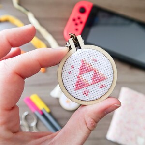 May include: A small, round cross-stitch with a pink and white triangle design, held in a wooden hoop. The design is a video game symbol. A Nintendo Switch console is visible in the background.