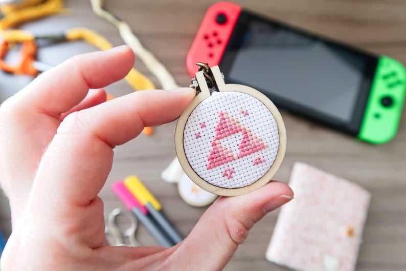 May include: A small, round cross-stitch with a pink and white triangle design, held in a wooden hoop. The design is a video game symbol. A Nintendo Switch console is visible in the background.