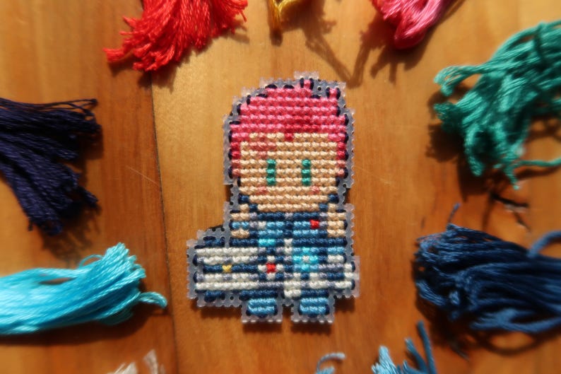 May include: A cross-stitch figure in a pixelated design, featuring pink hair, green eyes, and blue attire. The figure is surrounded by colourful embroidery floss in red, pink, teal, and blue, on a wooden surface.