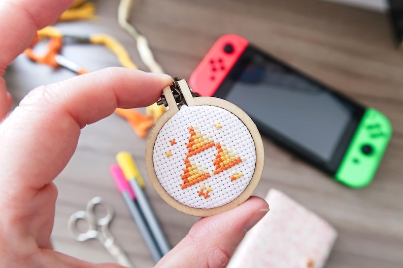 May include: A small, circular cross-stitch embroidery hoop featuring a geometric design in orange and yellow. The wooden hoop is held in a hand, with a Nintendo Switch console in the background. Craft supplies are also visible.