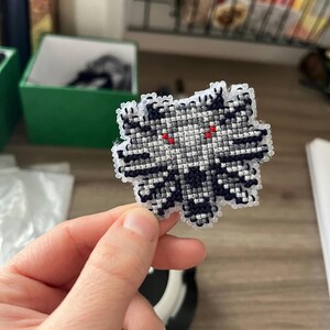 May include: A cross-stitch patch in the shape of a stylized wolf's head. The patch features a grayscale color scheme with black, white, and gray threads, and red eyes. The patch is held in a person's hand.