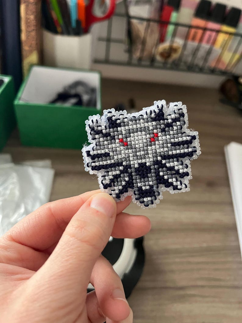 May include: A cross-stitch patch in the shape of a stylized wolf's head. The patch features a grayscale color scheme with black, white, and gray threads, and red eyes. The patch is held in a person's hand.