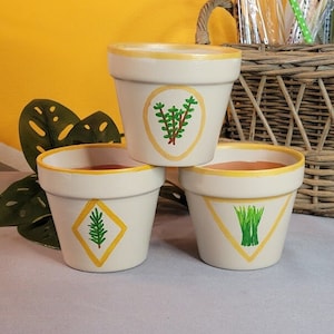 Puede incluir: Tres macetas de terracota blancas con bordes amarillos y hierbas verdes pintadas. Las macetas están apiladas una encima de la otra. Las hierbas son romero, tomillo y cebollino.