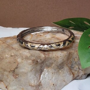 May include: A silver bangle bracelet with a gold and silver patterned design. The bracelet is on a rock with a green leaf in the background.