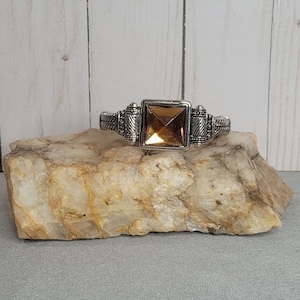 May include: A silver cuff bracelet with an ornate design and a large, square, amber-colored gemstone in the center.