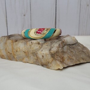 May include: A gold bangle bracelet with a white, turquoise, and red enamel design. The bracelet is decorated with clear rhinestones.