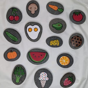 May include: Hand-painted rocks featuring various food items. The collection includes a cherry, mushroom, carrot, tomato, cucumber, fried egg, avocado, cheese, cookie, pumpkin, watermelon, broccoli, ice cream cone, orange slice, and strawberry.