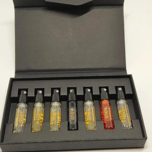 Op de afbeelding: Een zwarte doos met een scharnierend deksel, met zeven kleine parfumflesjes. De flesjes zijn van helder glas met zwarte sproeiers en etiketten. De vloeistoffen binnenin hebben verschillende kleuren, waaronder geel en rood.