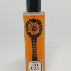 Könnte beinhalten: Eine durchsichtige Glasflasche mit einer orangefarbenen Flüssigkeit. Die Flasche hat einen schwarzen Verschluss und ein schwarzes Etikett mit dem Text "SIGILLI" und "VO LUM NA" in Weiß. Das Etikett zeigt auch ein schwarzes und goldenes Wappen mit dem Buchstaben "B" in der Mitte.