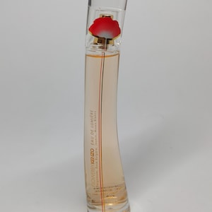 Op de afbeelding: Een heldere glazen parfumfles met een rood bloemdesign aan de bovenkant. De fles heeft een gebogen vorm en bevat een lichtoranje vloeistof. De tekst "Flower by Kenzo Eau de Lumiere" staat op de fles.