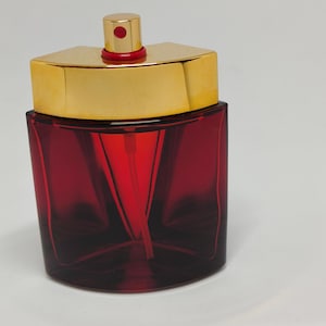 Könnte beinhalten: Eine rote Glasparfümflasche mit goldfarbenem Deckel und rotem Zerstäuber. Die Flasche hat eine rechteckige Form mit abgerundeten Kanten. Der Deckel ist rechteckig mit einem kleinen, runden Zerstäuber. Die Flasche steht auf weißem Hintergrund.