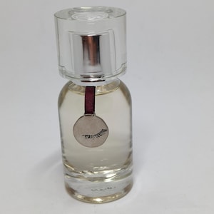 Può includere: Un flacone di vetro trasparente con un tappo argentato e un ciondolo argentato con un design di ciglia appeso a un nastro bordeaux. Il flacone è riempito con un liquido trasparente.