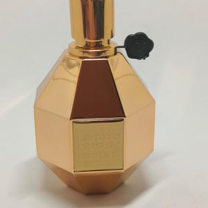 Puede incluir: Un frasco dorado de perfume Viktor & Rolf Flowerbomb Rose Explosion. El frasco tiene forma de gema multifacética y tiene una tapa negra con un emblema de rosa.