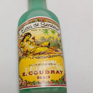 Puede incluir: Una botella de perfume vintage de vidrio verde claro con una etiqueta amarilla que presenta una mujer y dise&ntilde;os florales. La etiqueta dice "Eau de Senteur" y "E. Coudray, Paris". La botella tiene una tapa verde y probablemente es de Francia.